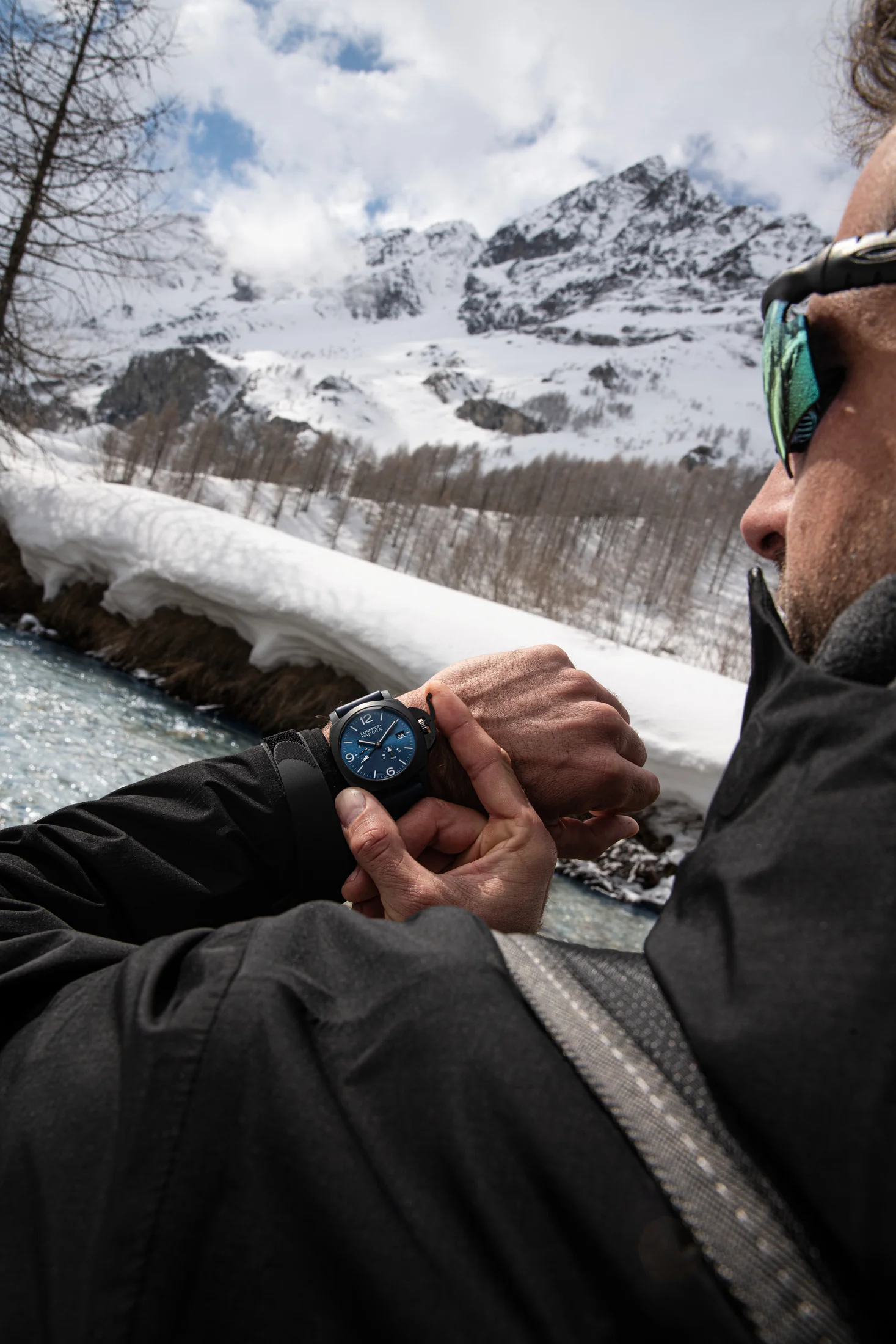 Explorer wearing a blue-dial Panerai watch beside a snowy mountain stream.